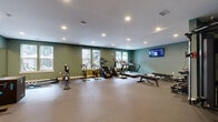 Located at 2001 Dabney Rd in Richmond, VA, this fitness center offers a well-appointed exercise room designed to support a full range of workout routines. The space is finished with durable rubber flooring throughout, providing a safe and practical surface for all types of training activities.

The cardio zone is lined with multiple treadmills, stationary bikes, an elliptical, and a stair climber, all positioned along windowed walls that allow natural light to filter in from multiple directions. A wall-mounted flat-screen TV is conveniently positioned for viewing during workouts.

The strength training area is equally comprehensive, featuring a tiered dumbbell rack stocked with a wide range of free weights, a functional cable machine, a power rack, and various benches for pressing and stretching exercises. An additional rack holds kettlebells and medicine balls, accommodating diverse training styles. A large wall mirror enhances the sense of space while allowing users to monitor their form.

The room is painted in a calming sage green, complemented by white trim and a clean white ceiling with recessed lighting. The overall layout is open and thoughtfully organized, ensuring users can move between equipment areas with ease. The combination of natural light, modern equipment, and a functional design creates an inviting and motivating environment for fitness.