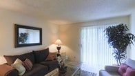 This single-floor space offers a comfortable and functional layout designed with everyday living in mind. The living area serves as the heart of the home, characterized by soft, neutral wall tones and a textured ceiling that adds subtle visual interest. A large sliding glass door fitted with vertical blinds allows natural light to fill the room while providing a connection to the outdoors. The space is thoughtfully arranged to accommodate relaxed seating, with room for a sofa, accent tables, and decorative touches that contribute to a warm, welcoming ambiance. A framed wall mirror enhances the sense of openness, reflecting light and adding depth to the room. The overall atmosphere is cozy yet bright, with a clean, understated aesthetic that lends itself well to personalization. The single-floor layout ensures ease of movement throughout the space, making it both practical and accessible. With its natural light, functional design, and inviting character, this space provides a comfortable setting suited to a variety of living arrangements.