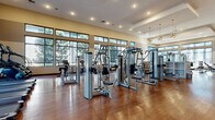 Located at 8555 Belle Dr in Highlands Ranch, CO, this expansive fitness center and yoga room offers a generously sized, single-floor facility designed to support a full range of exercise disciplines. The main exercise floor is outfitted with an extensive lineup of Cybex strength-training machines, cable systems, free weights, and cardio equipment including treadmills and ellipticals, all arranged across gleaming hardwood-style flooring. Large banks of windows line the perimeter walls, flooding the space with natural light and offering views of the surrounding landscape. A striking Rocky Mountain-themed photographic mural wraps across a prominent interior wall, reinforcing the Colorado aesthetic and adding visual depth to the environment. Recessed lighting and stylish pendant fixtures with globe-cluster designs illuminate the space warmly throughout. A dedicated group fitness and yoga room is framed by tall dark wood columns and floor-to-ceiling glass partitions, creating an airy, open-concept studio bathed in natural light from expansive windows. This studio accommodates spin bikes for cycling classes and open floor space for yoga and group workouts. The room is equipped with a large flat-screen display, a mini-refrigerator, yoga mat storage, and a colorful dumbbell rack. The overall ambiance is modern, energetic, and thoughtfully designed to deliver a premium fitness experience.