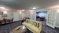 This single-floor community amenity space is thoughtfully designed for leisure and social interaction, offering a seamless blend of comfort and classic style. The layout flows between two interconnected areas joined by a set of elegant French doors with glass pane detailing, allowing an open yet defined separation between zones.

The main lounge area features richly patterned carpeting in blue and brown tones, crown molding throughout, and warm flush-mount ceiling lighting. Seating is generously arranged, including a light yellow sofa adorned with striped accent pillows, upholstered armchairs, and small side tables and round wooden accent tables, creating inviting conversational groupings. A large flat-screen television is mounted on a deep blue accent wall, complemented by a decorative media console below and framed nature-inspired artwork.

Through the French doors, a dedicated billiards room awaits, anchored by a full-size pool table and governed by a relaxed, club-like atmosphere. Deep blue walls, a ceiling fan, and a curated selection of framed vintage photographs lend the space a distinguished character. Bar-height chairs and a leather armchair provide casual seating along the walls. Together, the areas form a versatile, welcoming gathering space well-suited for both quiet relaxation and active recreation.