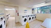 Located at 210 Steeles Ave W in Brampton, Ontario, this shared laundry facility is a well-equipped, single-floor space designed to serve building residents efficiently. The facility is divided into two laundry rooms, together offering a generous number of front-loading washing machines and stackable dryer units, providing ample capacity for simultaneous use. Light-toned ceramic floor tiles run throughout, contributing to a clean and practical environment.

One laundry room benefits from large windows that allow natural light to fill the space and provide views of the surrounding greenery, creating a more comfortable atmosphere. A utility sink and spacious folding tables are conveniently positioned for residents' use. The second laundry room houses additional stackable dryer units along both walls, maximizing machine availability within the footprint.

Adjacent to the laundry rooms is a dedicated waiting area, furnished with cushioned seating chairs and a table, offering residents a comfortable place to wait. A bulletin board displaying notices and informational postings is mounted on the wall, alongside a water dispenser for added convenience. Fluorescent ceiling lighting ensures consistent illumination throughout all areas. The overall layout is functional and thoughtfully organized, prioritizing ease of use and accessibility for all building residents.