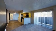 This single-floor residence offers a practical and open layout designed for comfortable everyday living. The living area features plush carpeting underfoot and is bathed in natural light streaming through a large sliding glass door dressed with vertical blinds, hinting at outdoor access beyond. A textured popcorn ceiling adds a classic touch throughout the space, while a ceiling fan with a light fixture provides both comfort and illumination.

The kitchen is positioned at the heart of the home, accessible via a raised pass-through counter that connects it to the living area. Warm wood-toned cabinetry lines the kitchen walls, complemented by black appliances including a range, microwave, and refrigerator. An overhead light fixture keeps the kitchen well-lit and functional.

Adjacent to the living space, a mirrored wall feature amplifies the sense of openness and reflects light throughout the room, adding a decorative and spacious quality. The flooring transitions from carpet in the living area to hard flooring in the kitchen, offering a clear and logical separation of spaces. Overall, the unit presents a cohesive, functional interior with classic residential finishes throughout.