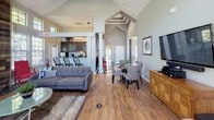Located at 1735 Mars Hill Dr. in Dayton, Ohio, this single-floor community space is designed to impress with its dramatic vaulted ceilings and open, airy layout. The main living area serves as the centerpiece of the space, featuring soaring cathedral ceilings with clerestory windows that flood the room with natural light and offer views of the surrounding greenery. A stunning floor-to-ceiling wood-planked accent wall frames a classic fireplace with a dark stone surround, creating a warm and inviting focal point. Rich hardwood flooring runs throughout, tying the open-concept layout together seamlessly. The adjacent dining area benefits from the same elevated ceilings and abundant window light, offering an intimate yet open atmosphere. The fully equipped kitchen features dark espresso cabinetry, stone-look countertops, a mosaic tile backsplash, stainless steel appliances, and a peninsula with bar seating, all illuminated by a modern geometric chandelier. A glass-paned door provides direct access to an outdoor patio area. Decorative archways with column details elegantly define transitions between zones. The space is bathed in natural light from multiple window banks and benefits from views of the outdoor pool area, making it a functional and welcoming environment for gathering and community activities.