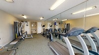 Located at 16211 Parkside Ln in Huntington Beach, California, this fitness center is a well-appointed exercise facility designed to serve the needs of its community. The room features a practical, carpet-covered floor and a textured ceiling with flush-mounted fluorescent lighting that ensures the space is well lit throughout.

The facility is outfitted with a diverse selection of cardio equipment, including multiple treadmills, elliptical trainers, and a recumbent bike, providing users with a variety of aerobic workout options. A multi-function strength training machine and a weight bench with free weights round out the offerings, catering to those focused on resistance training. A set of dumbbells is also available for supplemental exercises.

Full-length mirrors line one wall, visually expanding the space and allowing users to monitor their form during workouts. Wall-mounted fans provide additional air circulation throughout the room. A large sliding glass door and wide windows along one wall flood the interior with natural light and frame views of the lush, palm-lined grounds outside, creating an inviting and motivating workout environment.

Restroom facilities are accessible directly within the space, adding convenience for users. The overall atmosphere is clean, functional, and focused, making this fitness center a practical and accessible amenity for the Huntington Beach community it serves.