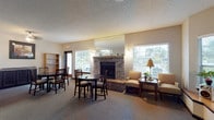 This single-floor communal space is thoughtfully designed to provide comfort and functionality for its occupants. The main gathering area serves as the heart of the space, anchored by a classic brick fireplace with a mirrored surround that reflects natural light throughout the room. Warm, neutral-toned carpet covers the floors, contributing to the inviting ambiance.

The lounge area features upholstered armchairs arranged around a side table, ideal for relaxed conversation. Adjacent to it, a dining area accommodates multiple guests with dark wood tables and ladder-back chairs. A striking bay window with a built-in wooden bench runs along one wall, bathing the space in natural light and offering views of the surrounding greenery. A television and audio equipment are also conveniently positioned in this area for entertainment.

The entry area includes a pair of glass-paneled double doors, a decorative ficus tree, and framed wall art, creating a welcoming transition into the main space. A dedicated kitchen area is equipped with stainless steel appliances, including a refrigerator, range, and over-the-range microwave, complemented by dark cabinetry and granite-style countertops — well-suited for preparing and serving meals or refreshments. Throughout, textured ceilings and ample window coverage with adjustable blinds complete the practical yet comfortable design.