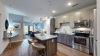 Located at 2439 Casona Ln in Melbourne, Florida, this single-floor residence offers a thoughtfully designed open-concept layout with contemporary finishes throughout. The kitchen serves as a central hub of the home, showcasing a granite-topped island with bar seating, two-tone cabinetry in warm wood and light gray tones, a white subway tile backsplash, and a full suite of stainless steel appliances including a refrigerator, range, microwave, and dishwasher. Pendant lighting above the island adds a modern touch, while wood-look flooring flows seamlessly into the adjacent dining and living areas. The living room is bright and welcoming, featuring a ceiling fan, a sliding glass door that opens to a private patio, and a cozy electric fireplace set within a rustic media console. The bedroom continues the coastal-inspired design with a bold blue accent wall, plush carpeting, and its own glass door providing additional outdoor access with natural light. The bathroom is well-appointed with a granite vanity, wood-toned cabinetry, a tub-shower combination, and wood-look flooring. Throughout the space, a cohesive blue-and-gray color palette creates a calm, sophisticated ambiance suited to Florida living.