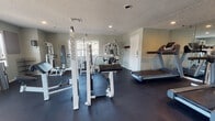 This fitness center is a dedicated, single-floor exercise facility designed to support a full range of workout routines. The space is finished with durable dark rubber flooring throughout, providing a safe and practical surface for all types of exercise. A textured ceiling with recessed lighting ensures the room is well-lit, while multiple windows along the walls allow natural light to fill the space and offer views of the surrounding outdoor area.

The cardio section is equipped with multiple treadmills and elliptical machines, giving users a variety of options for aerobic training. A large floor-to-ceiling mirror along one wall visually expands the space and allows users to monitor their form during workouts. Strength training equipment includes multi-function cable machines, weight benches, and additional resistance machines suited for both upper and lower body exercises.

The layout is thoughtfully organized, with cardio equipment positioned along the mirrored wall and strength machines distributed throughout the open floor area, allowing for comfortable movement between stations. A wall-mounted coat hook near the entry door provides convenient storage. The neutral gray walls contribute to a clean, focused atmosphere. Entry and exit are provided through clearly marked doors, including a dedicated exit with signage, ensuring safety and accessibility throughout the facility.