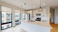 This contemporary single-floor residence showcases a clean, modern design throughout, with light-toned luxury vinyl plank flooring and crisp white walls creating a bright, cohesive aesthetic. The open-concept kitchen serves as the heart of the home, centered around a generous island with a quartz countertop and an undermount sink with a high-arc faucet. White shaker cabinetry with sleek matte black hardware lines the walls, complemented by a classic white subway tile backsplash. Stainless steel appliances — including a French door refrigerator, electric range, and built-in microwave — complete the well-equipped space. Geometric pendant lights add a stylish, industrial touch overhead, while large windows with roller shades allow abundant natural light to fill the kitchen and living areas. The spacious living area is bathed in light from a dramatic bank of floor-to-ceiling windows framing sweeping city views. Bedrooms are generously sized, with ceiling fans and ample closet storage featuring built-in shelving and hanging rods. The bathroom is finished with a white shaker vanity, quartz countertop, matte black fixtures, and a tub-shower combination with built-in shelving. An in-unit stacked washer and dryer adds everyday convenience to this thoughtfully designed residence.