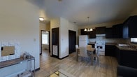 This single-floor space presents an open-concept layout that seamlessly connects the kitchen, dining, and living areas into one cohesive environment. The kitchen is outfitted with dark espresso cabinetry that contrasts sharply against light-colored walls, creating a bold visual statement. White appliances, including a refrigerator and electric range with overhead ventilation, contribute a clean, functional aesthetic. The countertops complement the darker tones of the cabinetry, adding to the kitchen's polished appearance.

Adjacent to the kitchen, the dining area is anchored by a light wood table paired with matching chairs, illuminated by an elegant chandelier with warm-toned bulbs that enhance the inviting atmosphere. The flooring throughout is a light-toned tile, lending a sense of continuity and spaciousness across the open floor plan.

A hallway branching from the main area leads to additional rooms, with dark-framed doors providing strong architectural contrast against the pale walls. Natural light filters through a window visible at the end of the corridor, brightening the space. The overall ambiance is clean, modern, and practical.