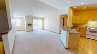 Located at 1702 Dublin Trail in Neenah, Wisconsin, this single-floor unit offers a practical and well-organized layout designed for comfortable everyday living. The open-concept living and dining area is generously sized, featuring soft wall-to-wall carpeting, a vaulted ceiling with a textured finish, and a sliding glass door fitted with vertical blinds that allows natural light to fill the space. A wall-mounted air conditioning unit provides climate control. The kitchen is positioned adjacent to the living area and is defined by a peninsula countertop with a built-in sink, warm maple cabinetry extending to the ceiling, neutral laminate countertops, tile flooring, and a full suite of appliances including an electric range with overhead ventilation and a refrigerator. The bedrooms are carpeted and bright, each featuring windows with blinds that bring in outdoor views of the surrounding trees. Ceiling fans are present in multiple bedrooms for added air circulation. One bedroom provides access to a utility and storage area. The bathroom features a wide vanity with ample cabinetry and drawer storage, a large wall mirror with overhead lighting, and tile flooring. Throughout the unit, warm wood-toned trim accents the neutral walls, contributing to a clean and cohesive aesthetic.
