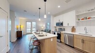 Located at 9 River St in Dover, NH, this thoughtfully designed one-bedroom unit offers a modern open-concept layout that maximizes every inch of its single-floor plan. The combined kitchen, dining, and living area serves as the heart of the home, anchored by a sleek center island with bar seating and stylish pendant lighting overhead. The kitchen is equipped with stainless steel appliances, two-tone cabinetry featuring white upper cabinets and warm wood lower cabinets, white tile backsplash, and quartz countertops. French doors in the living area flood the space with natural light and open to a view of the surrounding Dover cityscape. Wide-plank hardwood-style flooring runs throughout the main living areas, adding warmth and continuity. The bedroom is a calm, carpeted retreat with large windows that invite abundant natural light and offer exterior views. A full bathroom features a wood-toned vanity, soaking tub, and clean white tile finishes. The unit also includes in-unit laundry, a hallway connecting key areas, and generous closet storage — including a walk-in closet with wire shelving — providing practical everyday functionality alongside its contemporary aesthetic.