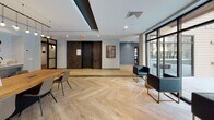 This thoughtfully designed single-floor amenity space combines modern aesthetics with a variety of functional zones, creating a versatile environment suited for both relaxation and social engagement. The layout is anchored by light-toned hardwood flooring in a herringbone pattern near the entrance, transitioning to wide-plank wood floors throughout the main lounge areas. Cove lighting integrated into tiered ceiling details adds a warm, layered ambiance across the space.

The primary lounge area features a large sectional sofa in steel blue upholstery, oriented around a stone-clad fireplace with a wall-mounted television above. Adjacent to this is a dedicated entertainment zone with a full-length shuffleboard table and raised banquette-style booth seating with high backs, offering a more private setting. A dark accent wall with curated artwork adds visual contrast to the otherwise light, neutral palette.

A demonstration kitchen anchors one section, complete with a long live-edge communal table, pendant lighting, a display wall of product samples, and countertop workspace with a sink. Floor-to-ceiling glazed doors and windows flood the space with natural light and provide direct views to the outdoors. Near the entrance, two leather accent chairs offer a welcoming seating area beside the elevator lobby.