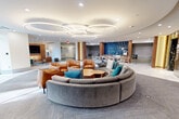 Located at 3400 Columbia Pike in Arlington, Virginia, this sophisticated lobby lounge offers a thoughtfully designed communal space that blends modern aesthetics with functional comfort. The centerpiece of the open floor plan is a large, curved sectional sofa in soft grey, accented with teal throw pillows and flanked by cognac leather armchairs and natural wood coffee tables — creating an inviting social hub. Overhead, a striking series of oversized circular ring pendant lights illuminate the space with a warm, contemporary glow against the smooth white ceiling.

The flooring transitions seamlessly between large-format marble-look tile and patterned carpet, delineating different zones throughout the lounge. A dedicated dining and café area features sleek bistro-style seating set against a bold blue herringbone tile accent wall, complemented by warm wood cabinetry and counters. A refined reception console with a decorative herringbone wood screen panel adds architectural interest near the entryway.

A more intimate seating alcove offers a large wall-mounted television framed in a warm wood surround with built-in backlit display niches, paired with a teal sofa, leather chair, and a dark oval coffee table — ideal for casual viewing or relaxed conversation. Recessed lighting throughout ensures consistent, clean illumination across every area of this well-appointed space.