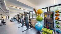 This fitness center offers a comprehensive workout environment housed within a space that blends industrial character with modern functionality. The layout spans a single floor in a long, open format, allowing for clearly defined zones dedicated to different types of training. Exposed timber beams and stone or concrete wall sections lend the space a distinctive architectural character, contrasting with the clean white walls and ceilings fitted with linear LED lighting that ensures consistent, even illumination throughout.

A row of Star Trac treadmills lines one side of the main floor, positioned in front of large, multi-pane windows framed in matte black. These windows allow abundant natural light to fill the space while providing views of surrounding greenery, creating an energizing atmosphere for cardio workouts. Alongside the treadmills, stationary bikes and elliptical machines further expand the cardio offerings.

The strength training area is well-stocked with barbells, weight plates, and dedicated racks, accompanied by a functional cable machine suitable for a wide variety of exercises. An accessories wall features resistance bands, stability balls in multiple sizes, and foam rollers, supporting flexibility and recovery routines. The durable, rubberized flooring throughout provides both safety and comfort. Overall, this fitness center is a thoroughly equipped facility designed to support a wide range of fitness goals.