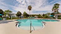Located at 3544 Saint Johns Bluff Rd S in Jacksonville, Florida, this outdoor amenity area offers a well-appointed resort-style pool experience. The centerpiece is a generously sized swimming pool with a curved, freeform shape, surrounded by a wide concrete pool deck that provides ample space for lounging and socializing. The pool reaches a depth of 4 feet 6 inches and is complemented by a separate hot tub/spa situated alongside rows of teal-colored lounge chairs, ideal for relaxation in the Florida sun.

A covered pavilion with a distinctive red tile roof borders one side of the pool area, offering shaded seating and shelter. Additional outdoor dining and seating areas are positioned throughout the deck, complete with umbrella-shaded tables in a vibrant turquoise tone that complements the pool's clear waters.

The space is richly landscaped with mature palm trees, fan palms, ornamental grasses, and lush tropical plantings that create a private, resort-like atmosphere. A perimeter fence with green space beyond adds a sense of openness while maintaining the area's defined boundaries. Pool rules signage is prominently displayed, noting dawn-to-dusk hours and community guidelines. The overall ambiance is bright, airy, and inviting, reflecting the warm Jacksonville climate.