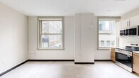 Located on the fifth floor at 929 Walnut St in the heart of Kansas City, MO, this efficiently designed studio unit offers a thoughtfully organized single-floor layout. The open living area is bathed in natural light from two large, classic double-hung windows that frame views of the surrounding downtown cityscape, creating a bright and airy atmosphere throughout.

The kitchen is a functional highlight of the space, featuring a clean, contemporary design with a mix of white upper cabinets and warm wood-toned lower cabinetry, accented by sleek black hardware. Stainless steel appliances — including a range, microwave, and dishwasher — are complemented by a full-size refrigerator, a single-basin sink, and a window that brings additional natural light into the cooking area.

The bathroom offers a minimalist aesthetic with a warm wood vanity, light countertop, modern black fixtures, and a combined shower and tub area with a curtain rod, all finished with neutral, easy-to-maintain surfaces.

Throughout the unit, light-toned flooring and crisp white walls with dark baseboards contribute to a clean, modern feel. An electrical panel and smart home control panel are neatly integrated near the entryway, adding practicality to the space's overall design.