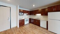 Located at 8412 22nd Ave S in Bloomington, Minnesota, this single-floor unit offers a thoughtfully organized layout that balances functionality with a clean, modern aesthetic. The kitchen serves as a central highlight, outfitted with rich dark-wood cabinetry extending to the upper walls, light countertops, a double-basin sink, and white appliances including a range, dishwasher, built-in microwave, and refrigerator. Recessed lighting illuminates the space efficiently, and wood-look plank flooring flows seamlessly from the kitchen into the open living area, creating a cohesive, airy feel throughout. The living space benefits from natural light admitted through a large window fitted with blinds, lending the room a bright and welcoming atmosphere. The bedroom areas feature plush carpeting underfoot and neutral wall tones, with one room offering a generous walk-in closet equipped with wire shelving and hanging rods along two walls, providing ample storage. The bathroom is finished in a clean, understated style with light tile flooring, a dark-wood vanity with a white countertop and single sink, a large mirror, and a shower-tub combination. Throughout the unit, consistent use of neutral tones and wood accents creates a cohesive and comfortable living environment.