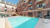 This residential community amenity space combines classic architectural design with refined interior details to create an inviting and functional environment for residents. The exterior showcases traditional red brick construction rising several stories, with private balconies and large grid-pane windows that reflect the building's timeless character. A generously sized outdoor swimming pool serves as a central outdoor feature, framed by a clean pool deck, shaded seating areas, and lush landscaping that enhance the overall resort-like atmosphere.

Inside, the clubhouse lounge is highlighted by a floor-to-ceiling stacked stone fireplace with a substantial wood mantel, creating a warm and inviting focal point. Comfortable seating arrangements, including plush sofas and wingback chairs in neutral tones, are thoughtfully positioned throughout the space. Tall ceilings amplify the sense of openness, while elegant chandelier lighting adds a sophisticated touch. Black-framed French doors and multi-pane glass panels connect interior spaces and allow natural light to flow freely throughout.

An additional lounge area features coordinated cream and soft blue furnishings alongside decorative accents, maintaining a cohesive, relaxed aesthetic. The entry area provides a welcoming arrival experience with soaring ceilings, tile flooring, and curated décor. Together, these spaces offer a polished and comfortable setting for community gatherings and everyday relaxation.