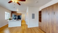 This single-floor residence offers a practical and cohesive layout designed for comfortable everyday living. Warm light hardwood flooring runs continuously throughout the space, creating a unified and inviting atmosphere across all rooms.

The open-concept kitchen is a functional highlight, featuring rich medium-toned wood cabinetry extending to the ceiling for generous storage. A full suite of black appliances — including an electric range, over-the-range microwave, and refrigerator — is complemented by a kitchen island with a double sink and light countertops. A window above the main kitchen area allows natural light to filter in while offering a view of the surrounding neighborhood.

The living area is bright and airy, benefiting from multiple windows and a ceiling fan with a light fixture that promotes air circulation and comfort. A glass-panel door opens directly to a private balcony, framing views of mature trees and providing seamless indoor-outdoor connectivity. An in-unit electrical panel and a built-in wood shelving unit with drawers add both practicality and storage versatility to the main living space.

A hallway connects the living and kitchen areas to the bathroom and additional rooms. The bathroom features light walls, hardwood-style flooring, and clean, simple fixtures. The overall design aesthetic throughout is clean and modern, with a neutral color palette that allows the warm wood tones to serve as the defining visual element.