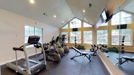 This fitness center offers a bright and energizing environment designed to support a well-rounded workout routine. The space is anchored by dramatic vaulted ceilings that soar overhead, creating an open, airy atmosphere that makes the room feel especially spacious. Large, multi-pane windows positioned along the front wall and sides allow abundant natural light to pour in, while also offering pleasant views of the surrounding greenery and outdoor areas. A ceiling fan is mounted at the peak of the vaulted ceiling, contributing to comfortable air circulation throughout the space. The flooring is covered in durable dark rubber gym matting, providing both traction and cushioning suitable for high-impact activity. The cardio section is well-appointed with multiple treadmills and elliptical trainers arranged in a row, while the strength training area includes a weight bench, a dumbbell rack with a full range of free weights, and additional resistance equipment. Wall-mounted televisions are positioned for easy viewing from the cardio machines. Recessed lighting complements the natural light, ensuring the space remains well-lit at all hours. The overall ambiance is clean, functional, and welcoming, making this fitness center a practical and motivating facility for daily exercise.