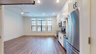 Located at 4220 Colony Plaza Dr in Charlotte, NC, this single-floor unit offers a thoughtfully designed open-concept layout that seamlessly connects the living area and kitchen. Rich hardwood-style flooring runs throughout the main living spaces, lending warmth and continuity to the interior. A bank of large windows floods the space with natural light, offering views of the surrounding neighborhood and creating an airy, welcoming atmosphere. The kitchen is outfitted with crisp white shaker cabinetry extending to the ceiling for ample storage, complemented by granite countertops, a herringbone-patterned tile backsplash, and a full suite of stainless steel appliances including a range, over-the-range microwave, dishwasher, and refrigerator. A distinctive sliding barn door with a matte black hardware rail adds a modern industrial accent and connects the main living area to the private spaces. The bathroom features light-toned tile flooring, a granite-topped vanity, and a glass-enclosed walk-in shower with subway tile surround. Recessed lighting and a ceiling fan enhance comfort throughout. The overall aesthetic is clean, contemporary, and move-in ready.