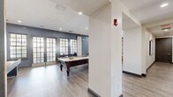 This thoughtfully designed single-floor amenity space offers a generous layout encompassing multiple living and gathering areas connected by well-proportioned hallways. The main game and lounge area serves as a centerpiece, featuring a full-size billiards table, a large flat-screen TV, and comfortable seating arranged on a patterned area rug. Grid-pane French doors flood the space with natural light and provide views of the surrounding greenery. A dedicated lounge area showcases a striking mixed-material accent wall of wood and concrete tiles, paired with plush golden leather chairs and a natural wood coffee table, creating a warm and inviting atmosphere. The kitchenette is fully equipped with dark gray shaker-style cabinetry, white quartz countertops, a sink, and a stainless steel mini-refrigerator, all framed by the same distinctive stone-and-wood accent wall. A private screening room offers tiered rows of cushioned armchairs, surround sound speakers, and projection screens for an immersive viewing experience. The covered outdoor patio extends the space with wicker seating and open views of the tree-lined surroundings. Throughout, light wood-style flooring, recessed lighting, and dark crown molding tie the design together cohesively.