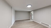 This single-floor unit has been thoughtfully updated with clean, modern finishes throughout. The main living area is open and airy, featuring light-toned vinyl plank flooring, neutral grey walls, crisp white baseboards, and a flush-mount ceiling light. A baseboard heater runs along one wall, and a doorway leads to the adjacent spaces, including a glimpse of a full-size refrigerator. The kitchen is equipped with stainless steel gas range, white cabinetry, a subway tile backsplash, granite-style countertops, and a window that brings in natural light and exterior views. The bedroom is carpeted in soft grey and features a large triple-window arrangement that fills the room with natural light, a baseboard heater, and a built-in closet with sliding doors offering practical storage. The bathroom is finished with large-format tile flooring laid in a staggered pattern, a full soaking tub with surround, and clean white fixtures. Throughout the unit, the consistent use of neutral tones, updated materials, and thoughtful detailing creates a cohesive and comfortable living environment.