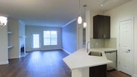 This single-story space showcases an open-concept layout that seamlessly connects the kitchen and living area. The kitchen is equipped with dark espresso cabinetry, a light-toned countertop with a built-in stainless steel sink, and a glass tile backsplash. Stylish pendant lights hang above the kitchen peninsula, complementing the track lighting overhead to provide layered illumination throughout. The living area features rich hardwood-style flooring that extends throughout the space, creating a warm and cohesive aesthetic. Crown molding adds a refined architectural detail along the ceiling. Large windows and a glass-paneled door allow generous natural light to fill the living area, offering views of the exterior. A hallway visible in the background suggests additional rooms or utility spaces beyond the main living area. The overall ambiance is clean, contemporary, and functional, with a neutral color palette accented by the contrast of dark cabinetry against light countertops and walls.