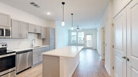 This single-floor residence offers a thoughtfully designed open-concept layout that seamlessly connects the kitchen, dining, and living areas. The kitchen is a standout feature, equipped with grey shaker-style cabinetry, a white subway tile backsplash, stainless steel appliances including a refrigerator with ice maker, electric range, microwave, and dishwasher. A large central island with a crisp white countertop provides additional workspace and serves as a natural gathering point. Pendant lighting above the island adds a modern touch, while light hardwood flooring flows throughout the main living areas.

The living area is bright and airy, benefiting from large windows and a glass-panel door that invite generous natural light and offer views of the surrounding neighborhood. A ceiling fan keeps the space comfortable year-round.

The bedroom is generously sized, featuring plush carpeting, a ceiling fan, and a large multi-pane window for natural light. A spacious walk-in closet with wire shelving provides ample storage.

The bathroom is clean and contemporary, featuring a wide vanity with quartz countertops, grey cabinetry, a large framed mirror, and a glass-enclosed walk-in shower with tiled walls. Light hardwood flooring completes the refined aesthetic of this well-finished residence.