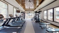 This single-story fitness center in Fort Mill, SC is thoughtfully designed to accommodate a wide range of workout styles in a spacious, open layout. The interior features a distinctive tray ceiling with warm wood paneling and recessed lighting, complemented by a large ceiling fan that enhances airflow throughout the space. Durable rubber and carpet-tile flooring defines separate workout zones, providing both safety and comfort underfoot.

The cardio section is lined with multiple treadmills and elliptical machines positioned to take advantage of natural light streaming through generously sized windows. A wall-mounted flat-screen TV keeps users entertained during workouts. The strength training area includes a multi-station cable machine, free weights, kettlebells, medicine balls, and a punching bag, offering comprehensive options for resistance training.

A standout feature is the full-panel glass garage door that opens entirely to the outdoors, seamlessly connecting the interior to an exterior pergola area and flooding the functional training zone with natural light and fresh air. This section also houses TRX suspension trainers and a wall-mounted storage rack. The overall ambiance is modern and energizing, with a clean, neutral color palette and abundant natural light throughout.