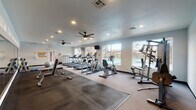 This fitness center offers a well-rounded exercise environment housed within a single-floor space. The room is finished with neutral gray walls, carpeted flooring, and rubberized mat sections strategically placed beneath weight and strength equipment to provide safety and durability. Recessed lighting throughout the ceiling, complemented by dual ceiling fans, ensures the space remains well-lit and comfortable during use.

Large windows line the perimeter walls, allowing abundant natural light to fill the room while offering views of an outdoor pool area, creating an open and inviting atmosphere. The cardio section is prominently featured, housing multiple treadmills, an elliptical machine, and a stationary recumbent bike, each positioned to take advantage of the outdoor views. Wall-mounted televisions provide entertainment options for cardio users.

The strength training area includes a multi-station cable machine and a dedicated free weight section equipped with a rack of dumbbells in varying sizes, exercise balls, and yoga mats. Workout reference charts are posted on the wall near the entry, offering exercise guidance. A small shelving unit provides additional storage. The overall layout promotes efficient circulation throughout the space, accommodating multiple users simultaneously with ease.