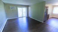 This single-floor space offers a clean, functional layout with a welcoming open feel. The main living area is finished with rich, dark hardwood-style flooring that contrasts elegantly with the soft sage green walls and crisp white baseboards, creating a calm, neutral palette throughout. A large sliding glass door serves as the focal point of the room, flooding the interior with natural light and providing direct access to an outdoor balcony with views of surrounding greenery. A wall-mounted air conditioning unit positioned near the door ensures year-round comfort. A interior door is visible along one wall, leading to additional areas of the unit. The partial view of a stainless steel refrigerator indicates that the kitchen is positioned adjacent to the main living space, allowing for a convenient, connected layout. The overall ambiance is bright, understated, and move-in ready, with modern finishes that provide a neutral foundation easily adaptable to a variety of interior styles.
