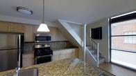 This two-floor space is thoughtfully designed to balance functionality and style. The kitchen serves as a central highlight, showcasing polished granite countertops with warm gold and beige tones that extend across both the island and main counter surfaces. Light wood cabinetry provides ample storage while lending a bright, clean aesthetic to the space. A granite tile backsplash complements the countertops and adds texture to the walls. Stainless steel appliances, including a gas range and an over-the-range microwave, offer a modern touch. A pendant light with a classic bell shade hangs above the kitchen area, providing focused task lighting alongside a flush-mount ceiling fixture. Adjacent to the kitchen, a staircase with a white-painted railing and wood handrail leads to the second floor, integrating seamlessly into the open layout. A window near the staircase landing allows natural light to filter into the transition area between floors. The overall design palette of neutral whites, light wood tones, and granite surfaces creates a cohesive and inviting atmosphere throughout the space.
