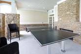 This covered outdoor amenity area is thoughtfully designed to blend rustic charm with functional recreation. The space is anchored by a striking floor-to-ceiling stacked stone fireplace, which serves as a natural focal point and provides warmth during cooler evenings. Complementing the fireplace, natural stone columns and archways frame open views to a resort-style swimming pool and outdoor lounge area beyond, creating a seamless indoor-outdoor connection.

The beadboard ceiling overhead adds a classic architectural detail, while recessed lighting ensures the space is well-lit throughout. White horizontal lap siding lines the perimeter walls, accented by a lower brick wainscoting detail that adds texture and visual depth. A set of elegant French doors with multi-pane glass inserts provides access to the interior, allowing natural light to flow between spaces.

Recreation is central to this area, with room for a ping-pong table alongside wicker high-top seating nestled between the stone columns — ideal for casual socializing. Additional seating is positioned near the arched openings, offering poolside views. A utility wall with a sink and bulletin board is also present, adding practical convenience. Together, the materials and layout create an inviting, community-oriented gathering space with a refined yet approachable character.