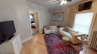 This single-floor home exudes classic charm through its thoughtful architectural details and warm, welcoming atmosphere. The living room serves as the heart of the space, showcasing beautiful hardwood floors that extend throughout and elegant crown molding that frames the ceiling. A ceiling fan provides both function and a traditional aesthetic touch overhead. Natural light filters in through windows, brightening the room and highlighting the soft, neutral gray walls that create a calm and airy backdrop. The living area is anchored by a vibrant jewel-toned area rug that adds a bold pop of color and personality, complementing the white upholstered seating arrangement centered around a circular glass-top coffee table. A framed world map adorns the wall, adding a curated, worldly accent to the space. Wide baseboards and clean trim work reinforce the home's classic character. An open doorway from the living room provides a seamless connection to the bedroom beyond, which continues the hardwood flooring and shares the same cohesive design language. The overall ambiance of the home is warm, well-maintained, and full of character, blending traditional architectural elements with comfortable, everyday livability.