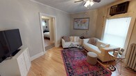 This single-floor home exudes classic charm through its thoughtful architectural details and warm, welcoming atmosphere. The living room serves as the heart of the space, showcasing beautiful hardwood floors that extend throughout and elegant crown molding that frames the ceiling. A ceiling fan provides both function and a traditional aesthetic touch overhead. Natural light filters in through windows, brightening the room and highlighting the soft, neutral gray walls that create a calm and airy backdrop. The living area is anchored by a vibrant jewel-toned area rug that adds a bold pop of color and personality, complementing the white upholstered seating arrangement centered around a circular glass-top coffee table. A framed world map adorns the wall, adding a curated, worldly accent to the space. Wide baseboards and clean trim work reinforce the home's classic character. An open doorway from the living room provides a seamless connection to the bedroom beyond, which continues the hardwood flooring and shares the same cohesive design language. The overall ambiance of the home is warm, well-maintained, and full of character, blending traditional architectural elements with comfortable, everyday livability.