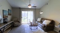 This single-floor residence offers a comfortable and well-appointed living environment designed for everyday ease. The living room serves as the heart of the space, characterized by neutral-toned walls and a textured ceiling that add depth and warmth to the room. A ceiling fan with a built-in light fixture is centrally positioned, providing both air circulation and ambient lighting. Natural light flows generously through a large sliding glass door dressed with white curtain panels, which opens directly onto an outdoor patio or balcony area, offering pleasant views of surrounding greenery. The living area is thoughtfully arranged with a leather sofa accented by plush throw pillows, a tufted accent chair, and a round wood-and-metal coffee table set atop a soft, layered area rug — creating a cozy and inviting seating arrangement. An arc floor lamp adds a stylish touch and supplemental lighting. The carpeted flooring runs throughout, contributing to the overall warmth of the space. The seamless connection between the interior living area and the outdoor space enhances the sense of openness, making this a comfortable and functional single-floor home.