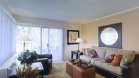 This single-floor residence features a thoughtfully designed living room that serves as the focal point of the space. Large sliding glass doors fitted with vertical blinds allow abundant natural light to fill the room, creating a bright and welcoming atmosphere while providing access to an outdoor area. The textured ceiling and warm, neutral wall tones establish a comfortable, understated backdrop throughout.

The living area is furnished with a spacious gray sofa adorned with decorative accent pillows in earth tones, complemented by a dark armchair positioned near the sliding doors. A pair of square brown leather ottomans double as a coffee table at the center of the room, adding both function and visual interest. A dark wood side table with a classic table lamp provides additional lighting and anchors one end of the seating arrangement.

Wall décor includes a framed artwork piece and a distinctive circular mirror set within a dark square frame, adding depth and character to the room. Plush, textured carpeting runs throughout the living area, contributing to the overall sense of comfort. The cohesive design elements come together to create a cozy yet modern living environment.