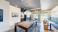 Located at 300 Highway 81 in McDonough, Georgia, this thoughtfully designed amenity lounge offers a dynamic and inviting environment for recreation and relaxation. The single-floor space is organized into several distinct activity zones that flow seamlessly into one another. At the heart of the room sits a full-size billiards table, positioned beneath a striking globe pendant light suspended from a coffered ceiling with a textured blue-gray finish and recessed lighting. Adjacent to the pool table is a lounge area featuring a bold blue channeled-back banquette set against an artistic botanical mural, accompanied by round ottomans and small bistro tables, creating an intimate social setting. A dedicated media lounge offers a sectional sofa, a wingback chair, and vibrant orange ottomans arranged around a wall-mounted display and a ceiling-mounted projector. Large windows fitted with roller shades allow natural light to fill the space while offering views of the surrounding greenery. An indoor putting green adds a recreational touch, while a kitchenette with navy cabinetry and a coffee station provides added convenience. Patterned carpeting and light hardwood flooring define the various zones, contributing to the space's modern and energetic aesthetic throughout.