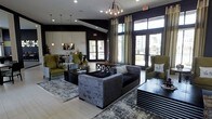 This single-floor space presents a sophisticated lounge environment defined by a bold, modern aesthetic. The interior is anchored by deep, dark-painted walls that create a dramatic contrast against the light tile flooring and large floor-to-ceiling windows and glass doors, which flood the space with natural light and offer views of the surrounding exterior. An ornate spherical chandelier serves as a central focal point overhead, complemented by pendant lighting near a secondary seating and counter area visible in the background.

The main seating area features a plush, grey crushed-velvet sofa paired with sleek, dark-finished coffee and side tables, creating a refined yet inviting arrangement. Olive green wingback chairs add a rich pop of color, grounding the space with warmth and contrast. A large, abstract area rug ties the groupings together cohesively. Tall, flowing curtains frame the expansive windows, adding elegance and texture. The background reveals an additional lounge nook with a mounted television, pendant lighting, and counter seating, suggesting a multifunctional layout suited for socializing or relaxation.