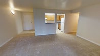 This single-floor residential unit offers a straightforward and functional layout designed to maximize livability. The main living area is generously sized, featuring neutral-toned wall-to-wall carpeting that creates a warm and inviting atmosphere. The space is bathed in a soft, even light that highlights the clean, understated design throughout.

A defining feature of the layout is the partial wall separating the living area from the kitchen, which includes a pass-through window that adds a sense of openness while maintaining a distinct division between spaces. The kitchen is well-appointed with warm wood-toned cabinetry providing ample storage, dark countertops, and standard appliances including a full-size refrigerator. The kitchen flooring transitions to a light-toned tile, offering a practical surface suited for cooking and meal preparation.

Adjacent to the main living space, an entryway with a white panel door provides access to an additional room, extending the functional footprint of the unit. The neutral gray walls throughout lend a clean, modern feel that serves as a versatile backdrop for a variety of interior styles. White baseboards and trim provide a crisp finishing touch, tying the overall aesthetic together cohesively.