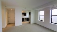 This single-floor unit features an open-concept layout that seamlessly connects the living area and kitchen. The living space is finished with neutral-toned carpeting and crisp white walls, creating a clean and airy atmosphere. A large window allows generous natural light to fill the room, offering elevated outdoor views that enhance the sense of openness.

The kitchen is visibly distinct with its dark espresso cabinetry, which provides a striking contrast against the white walls and light flooring. Stainless steel appliances, including a full-sized refrigerator and dishwasher, are integrated into the kitchen design. A sink with a standard faucet is positioned along the counter, and recessed lighting illuminates the kitchen workspace effectively.

The transition between the kitchen and living area is defined by a partial wall, providing visual separation while maintaining an open feel. The unit's neutral color palette, consistent flooring, and modern fixtures contribute to a functional and contemporary aesthetic throughout the space.