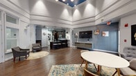 This single-floor sports lounge in Laurel, Maryland, offers a well-appointed, open-concept gathering space designed for both relaxed socializing and active entertainment. The standout architectural feature is the dramatic tray ceiling, accented with a deep blue inset and crown molding, adding height and visual interest to the main lounge area. Recessed and track lighting throughout ensures the space is well-illuminated at all times.

Rich hardwood flooring runs continuously across the entire space, grounding the open layout and complementing the cool blue-gray wall tones. The main lounge area is furnished with modern mid-century style seating, round tables, and layered area rugs, creating defined zones for conversation and dining. A large wall-mounted flat-screen television paired with a sleek media console serves as a central focal point for sports viewing.

Large white-framed French doors and transom windows along one wall flood the space with natural light and provide views to an adjacent outdoor area. A full kitchenette with dark espresso cabinetry, granite countertops, stainless steel appliances, and a bar counter with seating supports food and beverage service. Sports-themed artwork and decorative accents reinforce the lounge's identity throughout, completing a polished yet casual atmosphere.