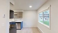 Located at 1000 W Ironwood Dr in Coeur d'Alene, Idaho, this thoughtfully renovated single-floor unit offers a clean, modern aesthetic throughout. Light wood-tone luxury vinyl plank flooring runs continuously across the entire space, creating a cohesive and bright interior.

The galley-style kitchen is a standout feature, equipped with stainless steel appliances including a range, microwave, refrigerator, and dishwasher. Bold navy blue lower cabinetry is paired with warm wood-toned upper cabinets and open shelving, all complemented by white quartz countertops and a undermount sink. A large window in the adjacent dining area fills the space with natural light and provides views of the surrounding community grounds.

The living area is anchored by a sleek built-in electric fireplace, adding warmth and a contemporary focal point to the room. A teal-painted Craftsman-style front door opens to views of the well-maintained community exterior, while additional windows ensure natural light reaches throughout.

The bedrooms feature neutral walls, ample natural light from windows with blinds, and wall-mounted heating and cooling units. Clean white panel doors and modern black hardware are consistent design elements throughout. A bathroom is conveniently accessible from the main hallway, completing this well-appointed, move-in-ready space.