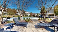 Located at 100 Riverview Pl in Jonesboro, Georgia, this thoughtfully designed outdoor amenity space offers a resort-style experience within a well-maintained residential community. At the heart of the area is a generously sized, freeform swimming pool with crystal-clear blue water, accessible via stainless steel entry rails and surrounded by a broad concrete sundeck. The deck is outfitted with sleek lounge chairs and seating areas, providing ample space for relaxation under the open Georgia sky. Adjacent to the pool, a dedicated outdoor dining area features table-and-chair sets, ideal for social gatherings. A charming single-story clubhouse with a covered porch and white trim anchors one end of the space, offering a welcoming focal point for residents. The surrounding landscape is meticulously maintained, showcasing manicured shrubs, ornamental trees, and winding concrete pathways that invite leisurely strolls through the grounds. Mature crape myrtles and towering evergreens provide natural shade and seasonal color throughout. Benches and small accent tables are thoughtfully placed along the walkways, creating quiet spots to enjoy the outdoors. The overall ambiance is serene and well-appointed, reflecting a strong attention to detail in both design and upkeep.