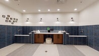 Located at 200 E Cama St in Charlotte, NC, this single-floor pet grooming facility is purpose-built to provide a clean, functional, and welcoming environment for pet care. The space is finished with a cohesive design palette centered around bold navy blue subway tile that lines the walls at mid-height, complemented by crisp white upper walls and a combination of large-format white floor tiles and classic penny mosaic tile underfoot.

The primary grooming area features multiple elevated pet bathing stations with white quartz-style countertops and warm wood-toned cabinetry below, providing ample storage for grooming supplies. Industrial-style wall sconces cast focused, warm lighting across the workstations, while recessed ceiling lights ensure the entire space is well-illuminated. Rolled fresh towels and grooming essentials are neatly arranged throughout, emphasizing the facility's attention to cleanliness and organization.

A dedicated steam room enclosed by frosted glass double doors offers an additional bathing or treatment option. The open floor plan allows for easy movement throughout, with floor drains integrated into the tile work for efficient cleanup. Playful, oversized dog portrait artwork adorns the walls, adding a welcoming and lighthearted character to the space. Leash hooks are conveniently mounted at multiple points, making the facility both practical and pet-friendly.