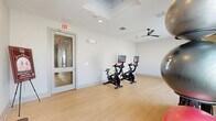 Located at 601 W Bacon St in Richmond, Virginia, this dedicated fitness room offers a well-appointed space for exercise and wellness. The room features light hardwood-style flooring throughout, contributing to a clean, modern aesthetic that is both functional and visually appealing. Bright white and neutral-toned walls keep the space feeling open and airy, while recessed lighting and a ceiling fan ensure comfortable conditions during workouts.

The fitness area is equipped with multiple stationary spin bikes fitted with interactive screens, making them suitable for structured cycling sessions. A storage rack holds an assortment of stability and medicine balls, while a colorful dumbbell rack provides a range of free weights for strength training. A simple bench offers a versatile surface for stretching or supplementary exercises. Framed artwork on the walls adds a touch of refinement to the utilitarian space.

Natural light filters in through a large window, brightening the room and creating an inviting workout environment. A glass-paneled exit door connects the fitness room to adjacent building spaces, ensuring convenient access. The open layout allows for flexible use of the floor space, accommodating a variety of exercise routines beyond the provided equipment. Overall, this fitness room delivers a practical and well-designed amenity for health-conscious users.