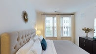 The bedroom is designed with a clean, contemporary aesthetic that emphasizes comfort and natural light. A generously sized tufted upholstered headboard in a soft neutral tone serves as the room's focal point, dressed with crisp white bedding and accented by a rich teal throw pillow that adds a subtle pop of color. Three large windows dressed with white horizontal blinds and flowing sheer cream curtains line the wall opposite the bed, allowing abundant natural light to fill the space while offering views of the surrounding buildings and greenery outside. The walls are finished in a soft, warm white that enhances the room's airy feel. A decorative sunburst mirror in antique gold adorns one wall, adding a stylish, artistic touch to the otherwise understated décor. A compact bedside table with a warm-toned lamp provides functional yet tasteful lighting. The ceiling is smooth and well-proportioned, contributing to the room's sense of openness. Overall, the bedroom conveys a serene, inviting atmosphere suited to rest and relaxation, balancing modern design sensibility with approachable warmth.