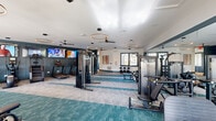Located at 1001 NW Cernan Dr in Huntsville, Alabama, this well-appointed fitness center offers a generously sized, single-floor exercise space designed to support a full range of workout routines. The interior is defined by a clean, contemporary aesthetic, featuring two-tone teal and gray patterned carpet flooring that adds visual depth and delineates functional zones throughout the space.

The facility is outfitted with a diverse selection of equipment, including treadmills, elliptical machines, stationary and spin bikes, a stair climber, cable and multi-use strength machines, and a dedicated free weight area with dumbbell racks and adjustable benches. Large wall-mounted flat-screen televisions are positioned throughout, providing entertainment during workouts.

Stylish circular pendant lighting and ceiling fans are suspended from the white ceiling, ensuring the space remains bright and comfortable. Full-length mirrors line portions of the walls, visually expanding the room and allowing users to monitor their form. Natural light filters in through multiple windows with teal-trimmed frames, complementing the facility's modern color palette. Colorful abstract artwork accents the walls, adding a creative touch to the energetic environment. The overall ambiance is clean, modern, and motivating—an ideal setting for a comprehensive fitness experience.
