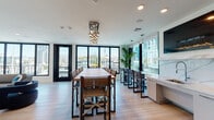 Located at 1331 N Cahuenga Blvd in the heart of Hollywood, this contemporary event and lounge space offers a sophisticated environment designed for gatherings and entertaining. The open floor plan connects a spacious lounge area with a large dining space, both bathed in natural light from expansive floor-to-ceiling black-framed windows that frame sweeping views of the Los Angeles skyline and surrounding hills.

The lounge area features light wood-style flooring, recessed lighting, and a wall-mounted flat-screen display, complemented by a generous L-shaped leather sectional sofa, a velvet accent chair, and a central coffee table — creating an inviting and relaxed atmosphere. Lush tropical plants are positioned throughout, adding a vibrant, natural touch to the modern aesthetic.

The dining area is anchored by a long communal wooden table with director-style chairs and a striking multi-globe pendant chandelier overhead. Adjacent bar-height seating along the window line provides additional space with direct views of the outdoor terrace and cityscape beyond.

The fully equipped kitchen showcases high-gloss grey cabinetry, a dramatic marble-veined stone backsplash with under-cabinet lighting, stainless steel appliances including a gas range and French-door refrigerator, and ample counter space. Glass-paneled doors add a refined architectural detail throughout the space.